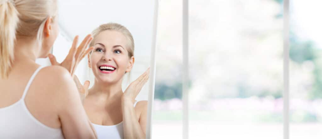 Mature woman looking happily at herself in the mirror after seeing fat transfer results